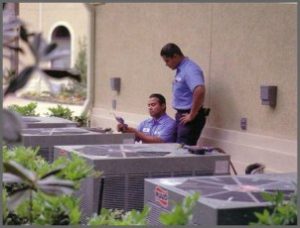 Two technicians in blue uniforms inspecting outdoor air conditioning units beside a building, with green foliage nearby, ensuring quality AC maintenance.